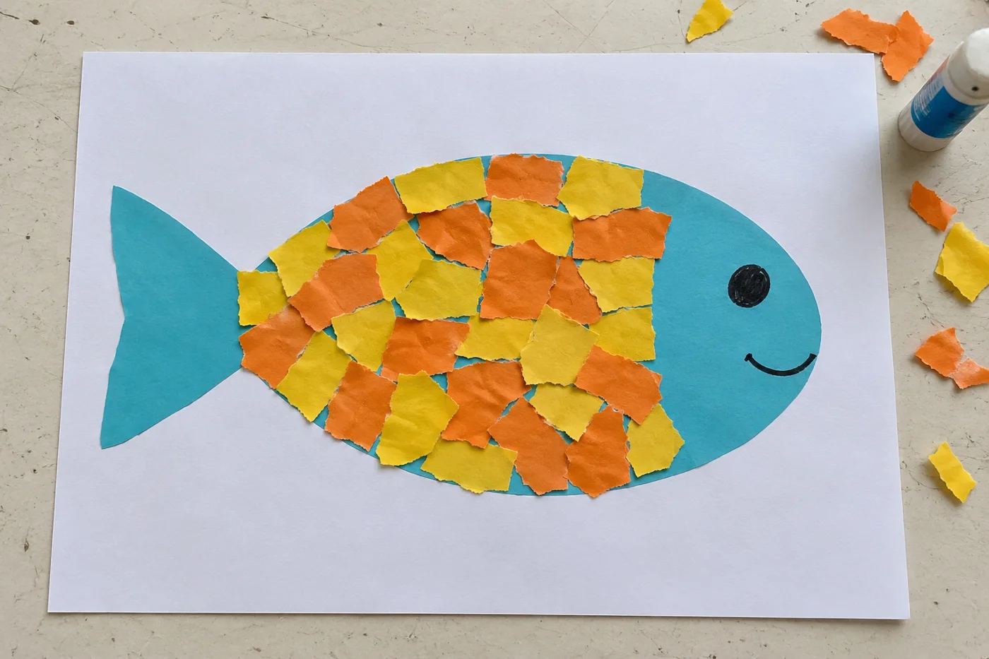 Poisson bleu souriant décoré de morceaux jaunes et orange en papier déchiré, sur une feuille blanche vue de haut