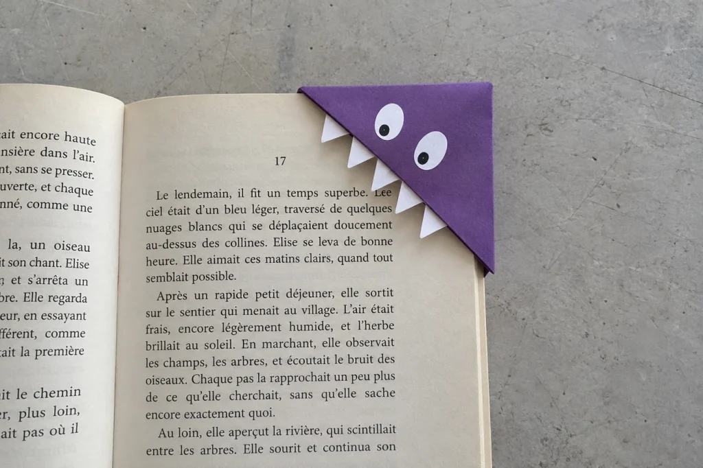 Marque-page monstre violet à deux yeux et cinq dents posé sur le coin d’une page, vu de haut