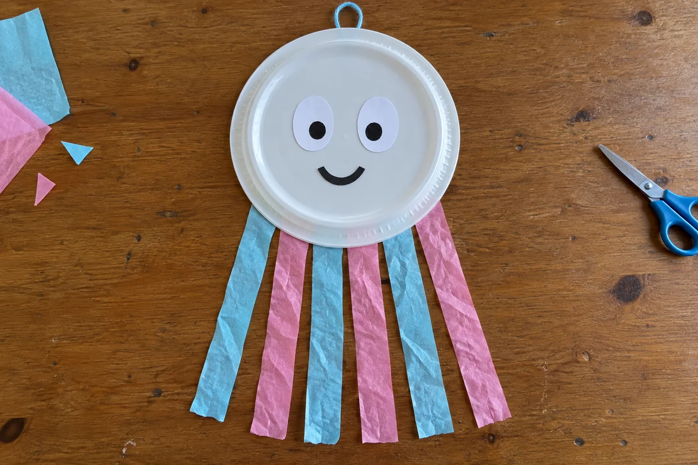Méduse souriante fabriquée avec une assiette blanche et six tentacules de papier bleu et rose, vue de haut
