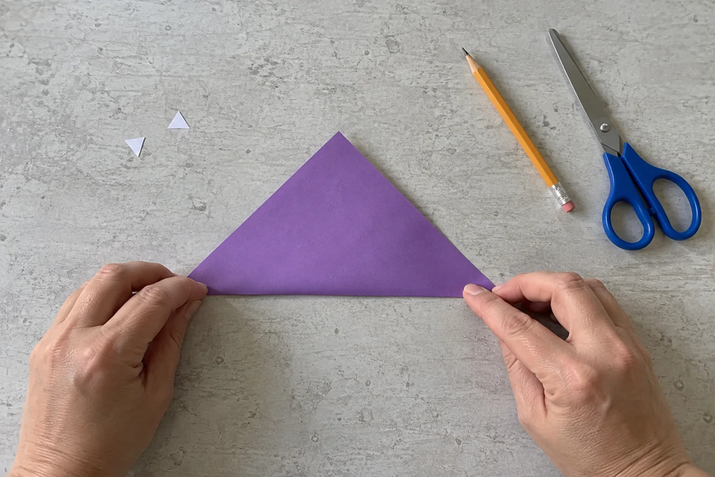 Mains alignant les coins d’un carré de papier violet plié en triangle sur une table grise, vue de haut